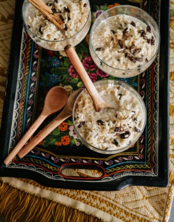 overhead shot of 3 footed glass goblets of arroz con leche topped with raisins and garnished with cinnamon sticks on a hand-painted serving tray.