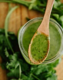 overhead shot of a glass mason jar filled with parsley cilantro chimichurri sauce with a wooden spoonful of it in focus and the jar and fresh herbs out of focus below it.