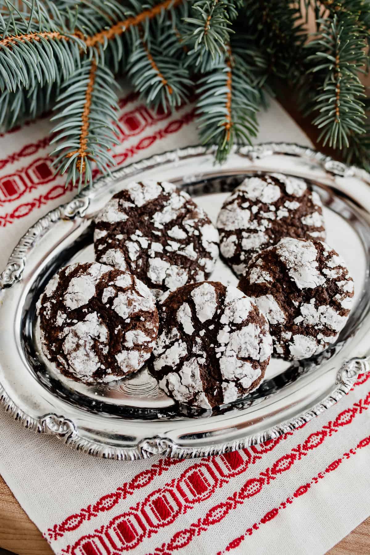 Mexican chocolate crinkle cookies on an oval silver platter on top of a red and white tea towel with a pine branch.
