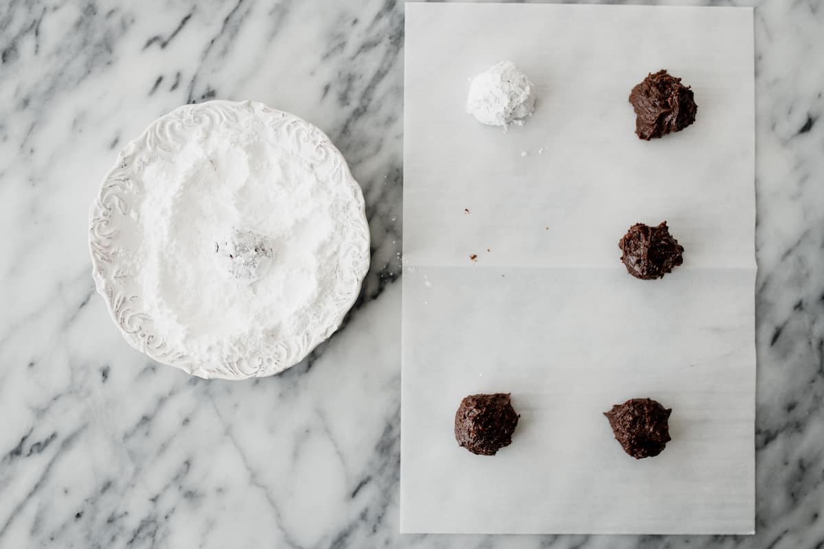 sheet of parchment with rolled cookie dough balls, one of which has been coated in powdered sugar.