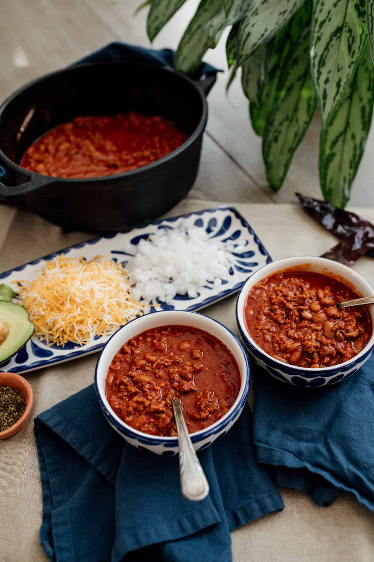 two blue and white bowls filled with chili con carne with a matching rectangular plate with onions, cheese and avocado as toppings.