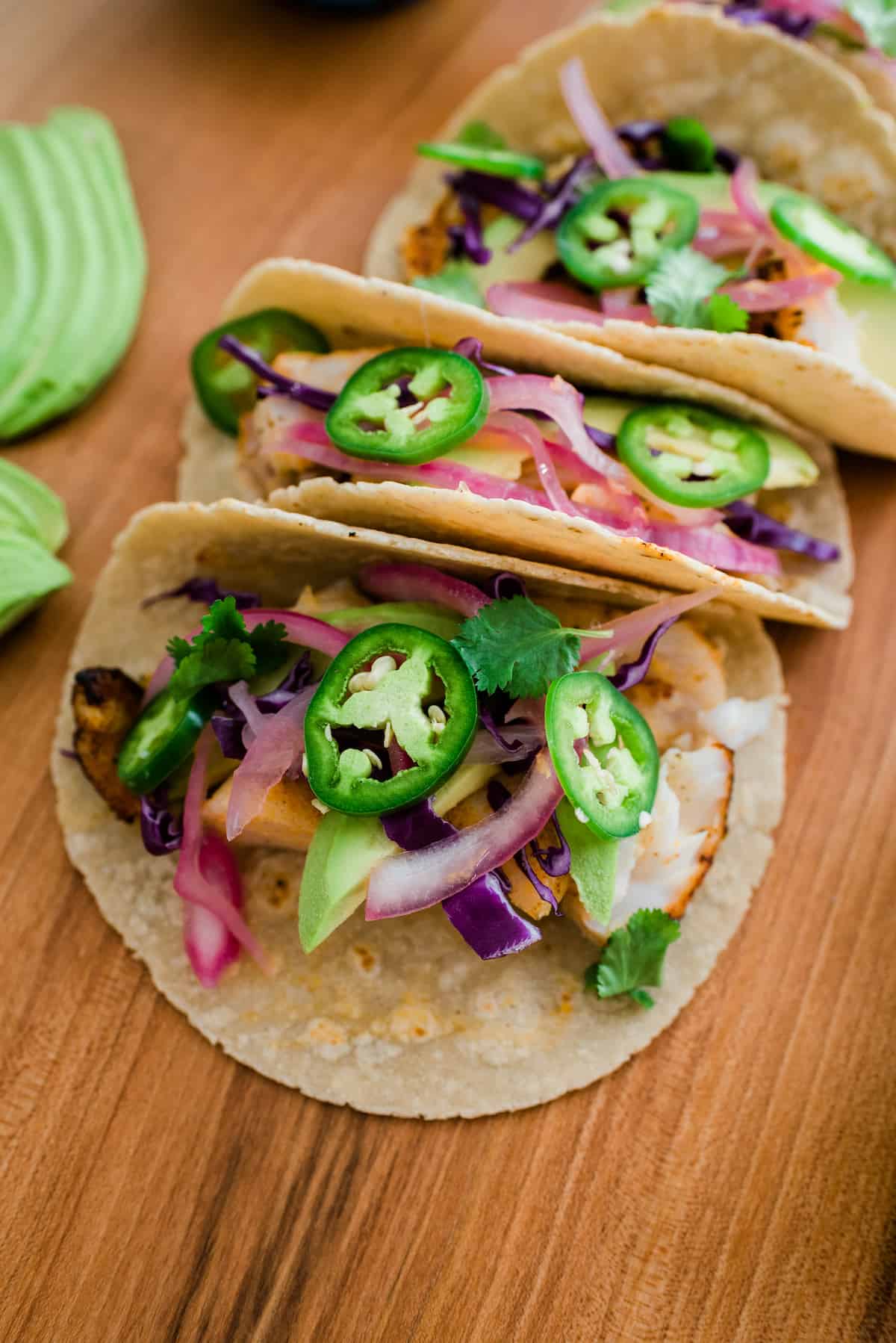 3 Easy Tilapia Fish Tacos served on a wooden table topped with picked red onions and fresh sliced jalapenos.