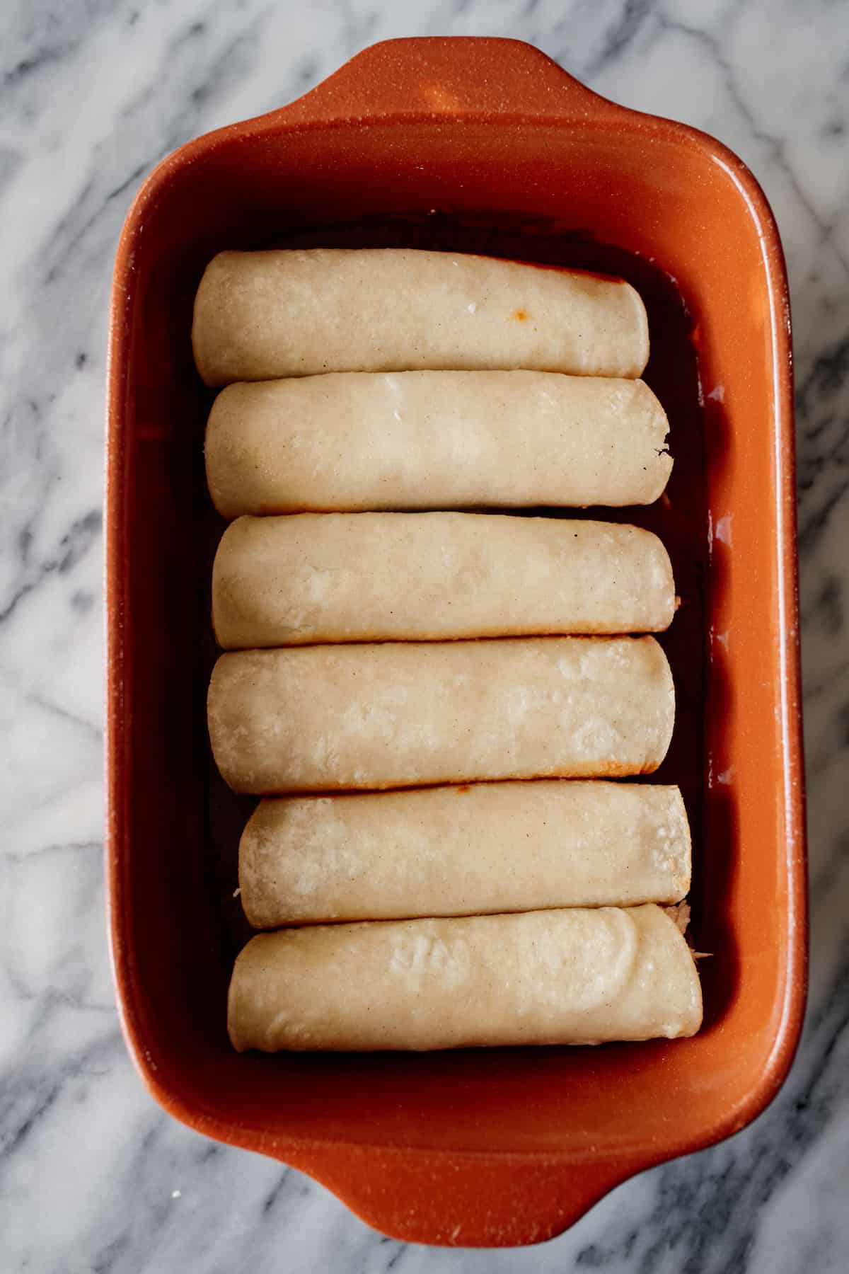 6 homemade easy chicken enchiladas in a terra cotta 9 x 13 baking dish before being covered with enchilada sauce and baked.