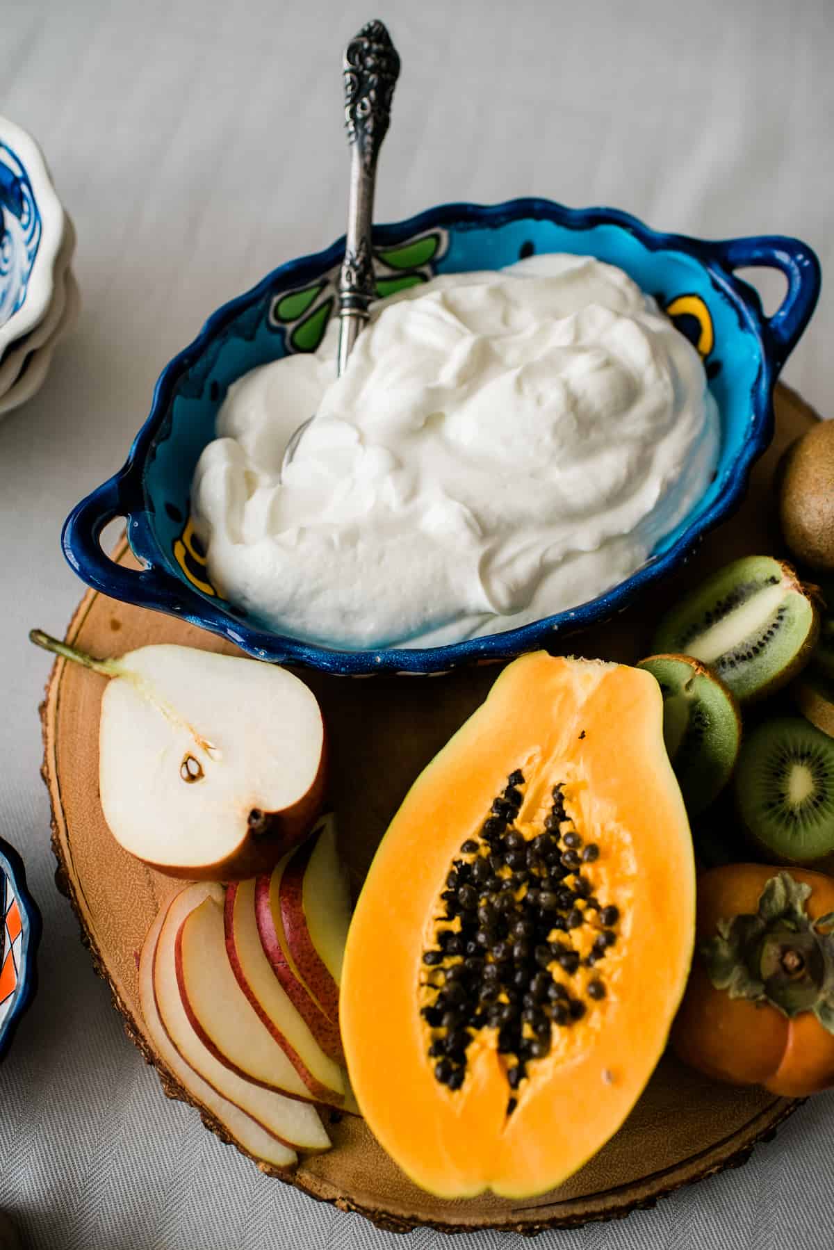 big blue serving bowl of yogurt on a wooden cutting board with an assortment of sliced fresh fruits for a DIY yogurt bar.