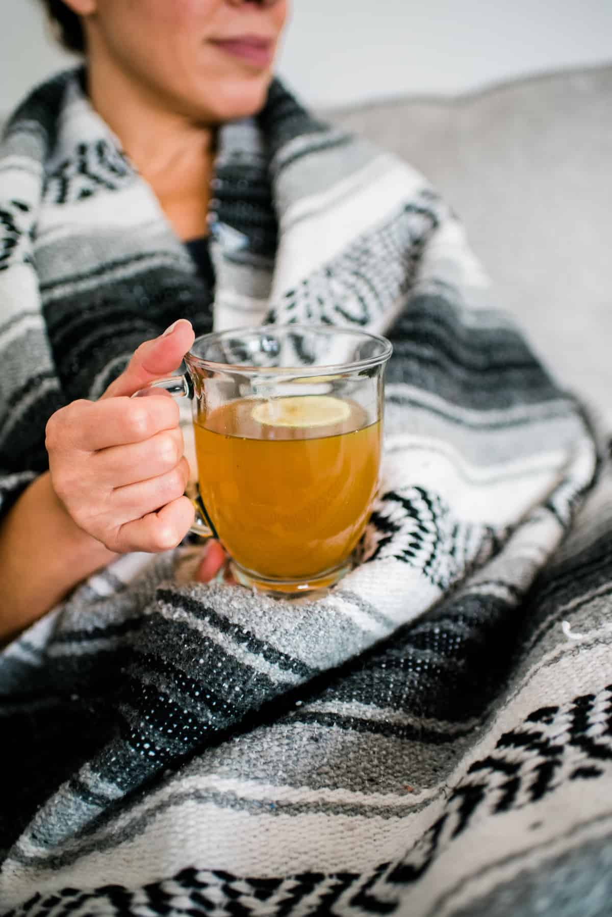 Latina woman wrapped in a grey, white, and black striped blanket holding a mug of Mexican oregano tea with a slice of lime.