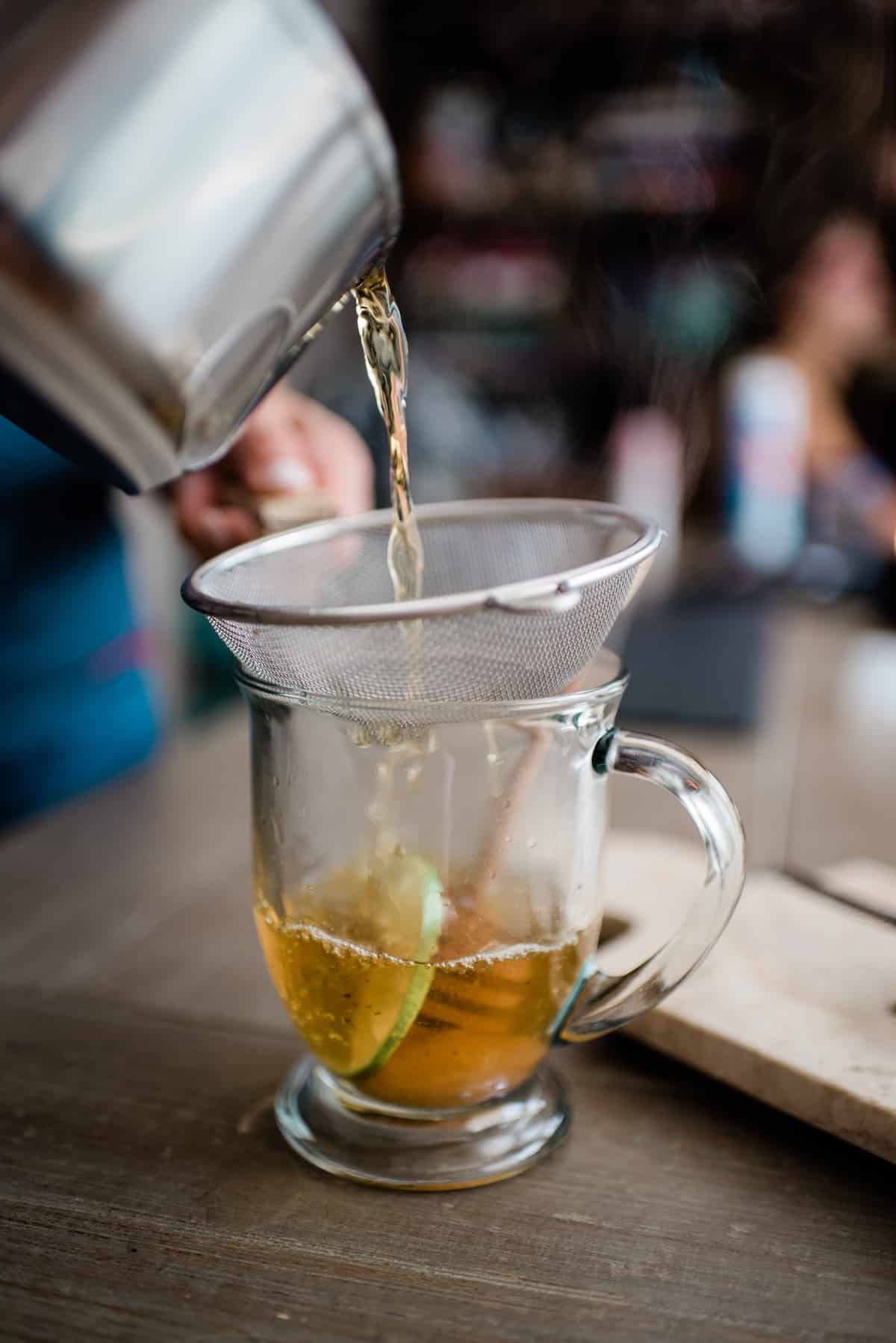 que muestra cómo colar el té de orégano de una cacerola a través de un colador de malla en una taza.