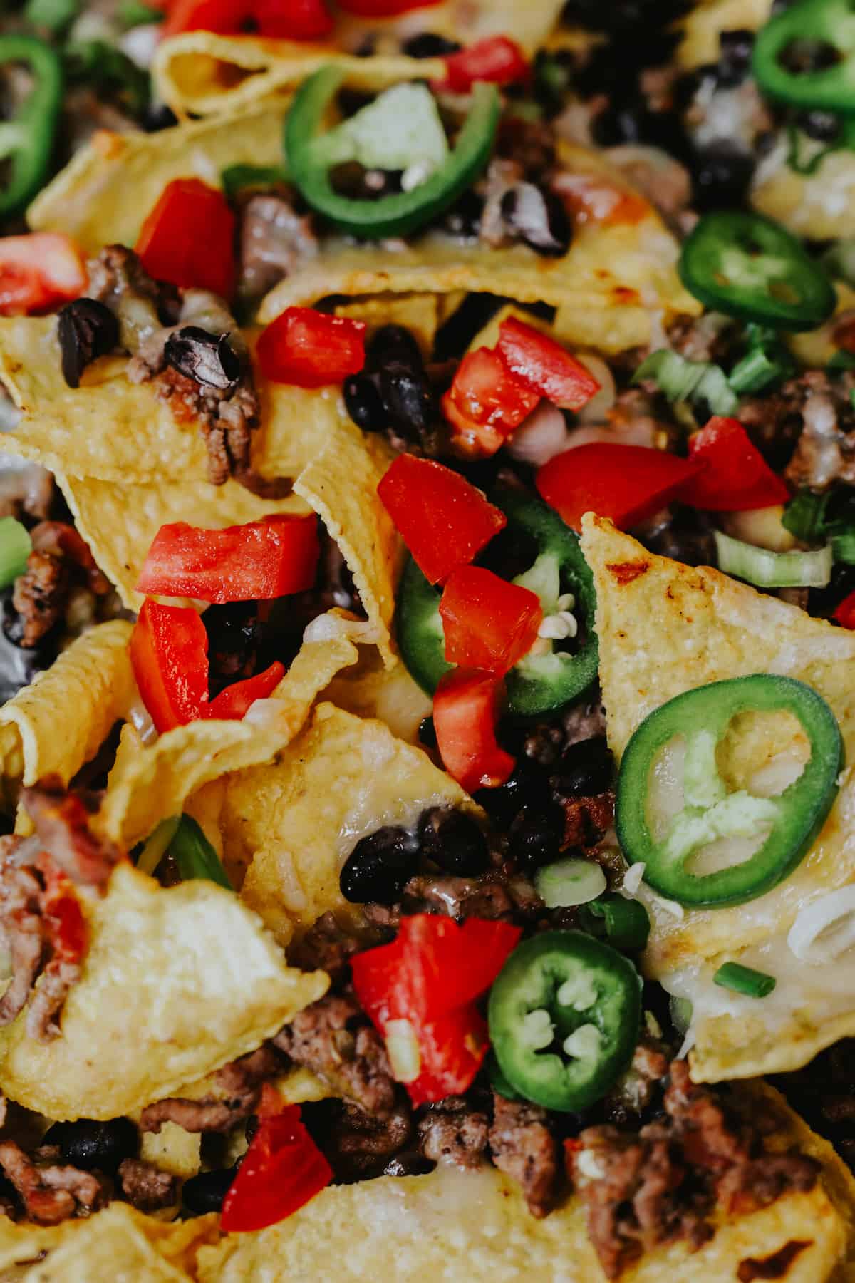 close up shot of classic baked sheet pan beef nachos topped with sliced jalapenos and diced tomatoes.