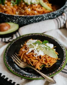 45 degree angle shot of a black plate with a green rim filled with a serving of fideo seco topped with avocado slices, a drizzle of crema, and a sprinkle of queso fresco.