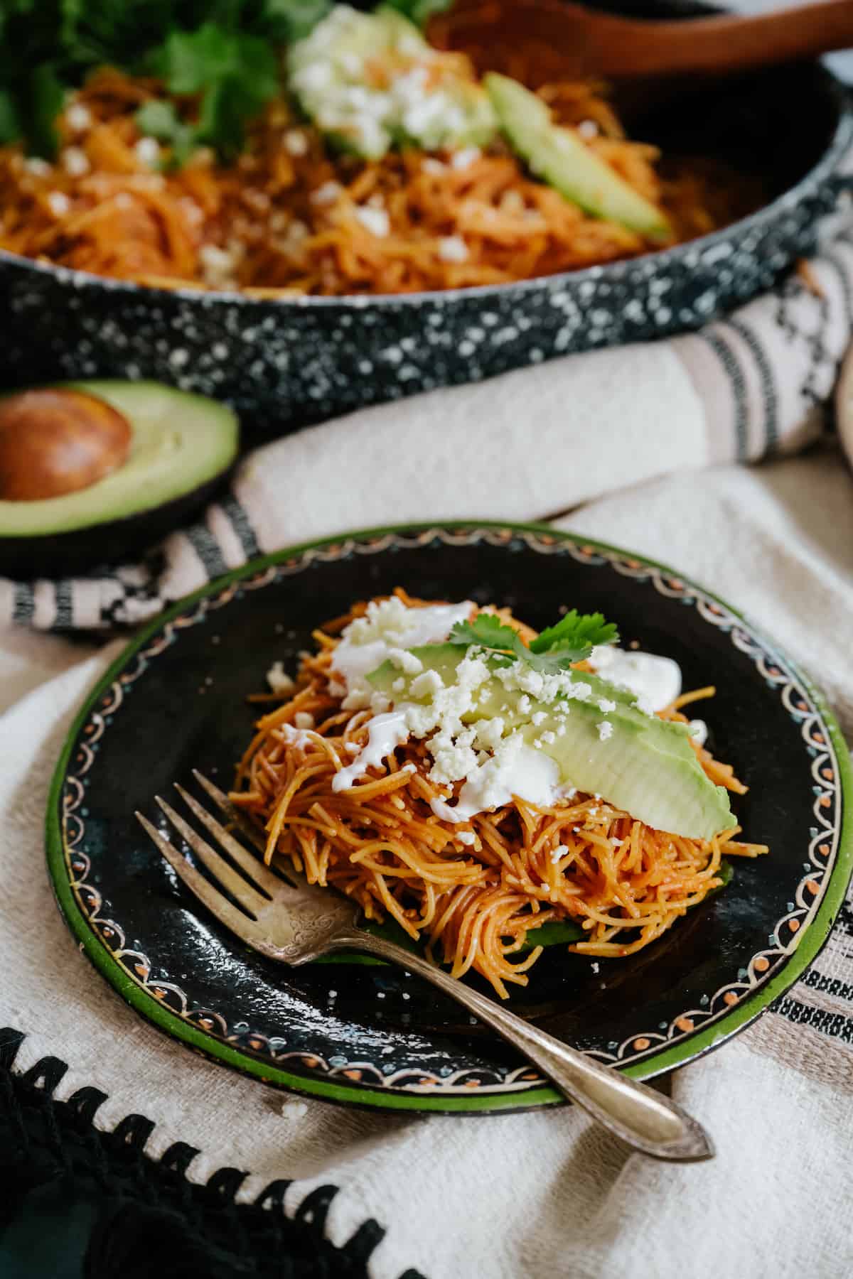 45 degree angle shot of a black plate with a green rim filled with a serving of fideo seco topped with avocado slices, a drizzle of crema, and a sprinkle of queso fresco. 