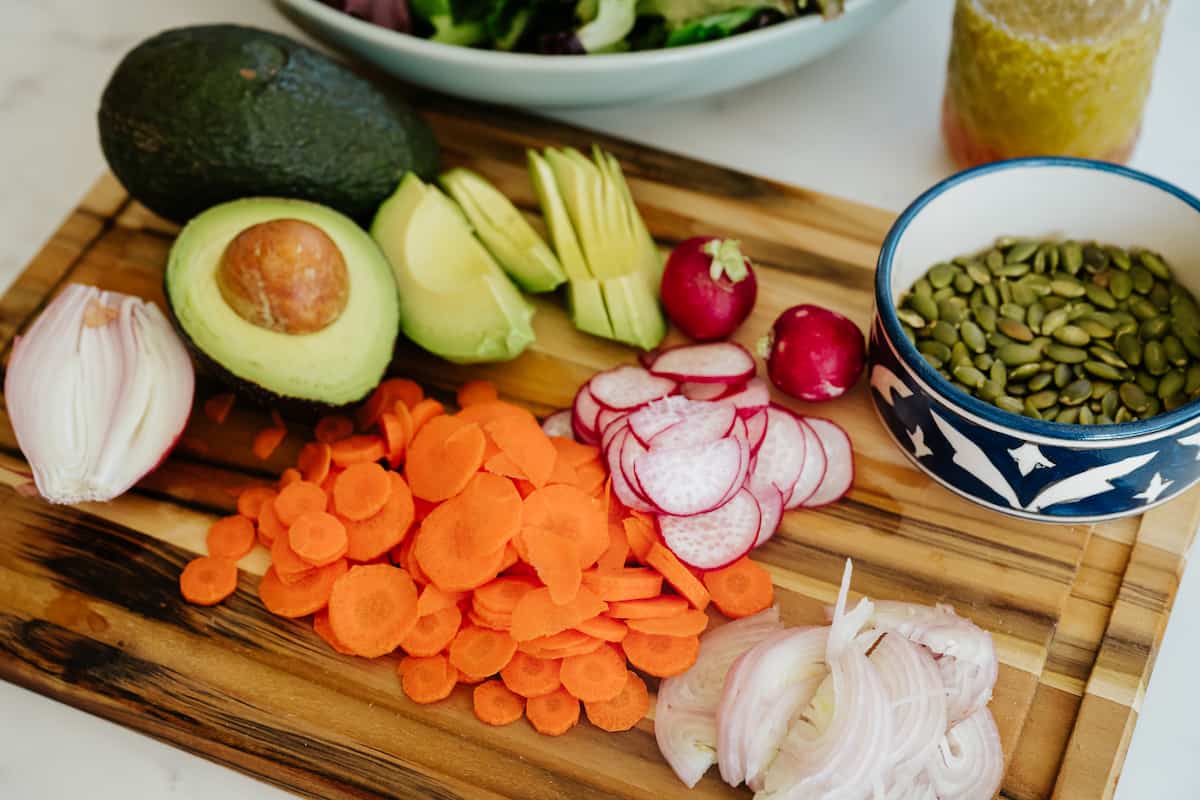 add-in ingredients for salad including thinly sliced shallots, radish, carrots, avocado and pepitas on a wooden cutting board.