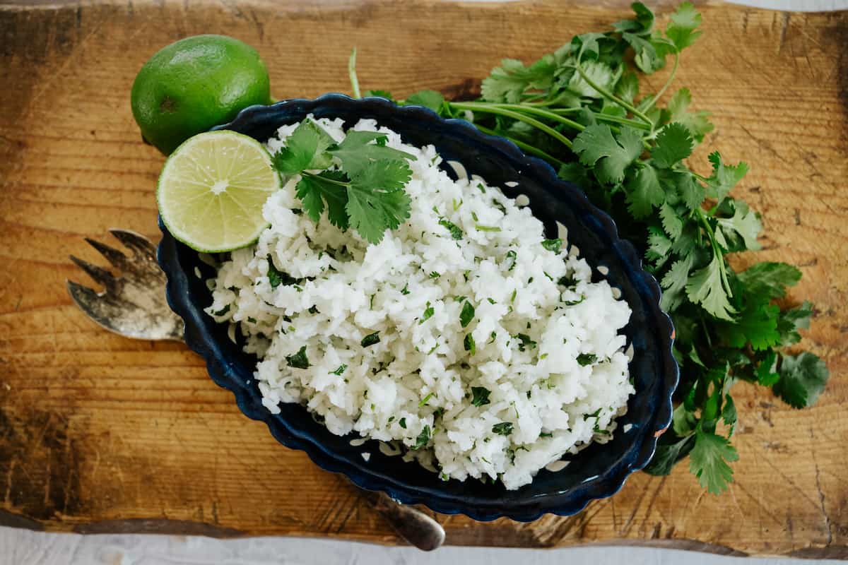 overhead shot of a serving bowl filled with Chipotle copycat cilantro lime rice on a wooden cutting board with a bunch of cilantro and an antique silver serving spoon on the side.