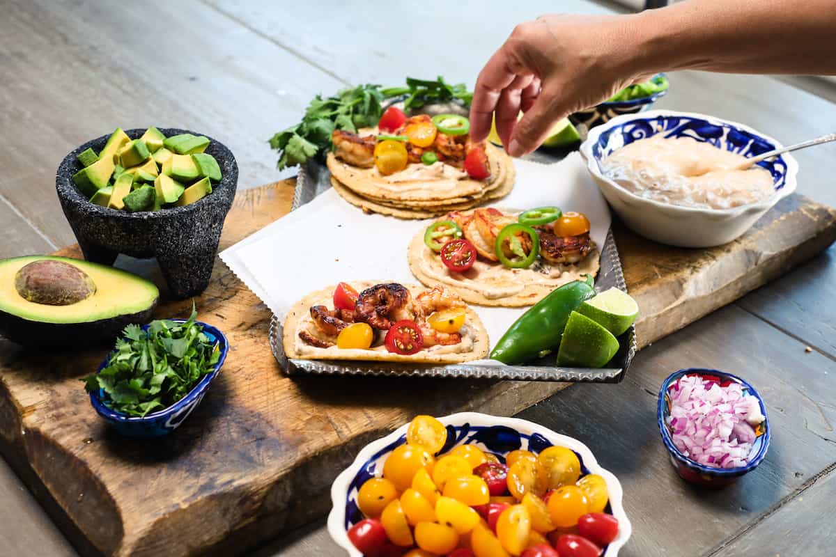Hand placing toppings and garnishes on shrimp tacos on a silver tray with bowls of colorful toppings.